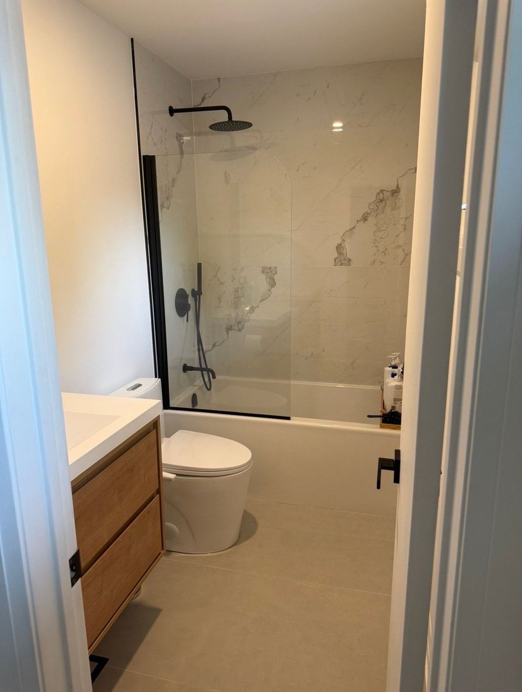 Bathtub with a hinged black-framed glass splash panel, large marble-look wall tile, matte black rain shower and hand shower, and a floating oak vanity, by NewGen Renovations Vaughan